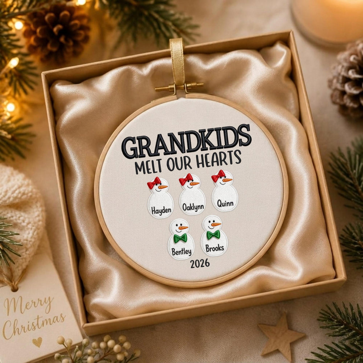 May include: A round wooden embroidery hoop ornament featuring white fabric with the black embroidered text GRANDKIDS MELT OUR HEARTS. Five snowman figures are displayed with names Hayden, Oaklynn, Quinn, Bentley, and Brooks, plus the year 2026.