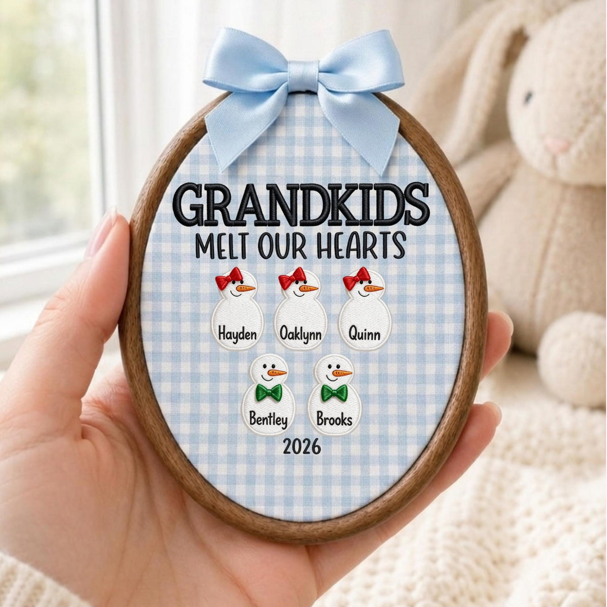 May include: An oval wooden embroidery hoop ornament featuring a light blue and white gingham fabric background. The text reads "GRANDKIDS MELT OUR HEARTS" above five embroidered snowman figures with names "Hayden", "Oaklynn", "Quinn", "Bentley", "Brooks" and "2026". A light blue satin bow is attached to the top.