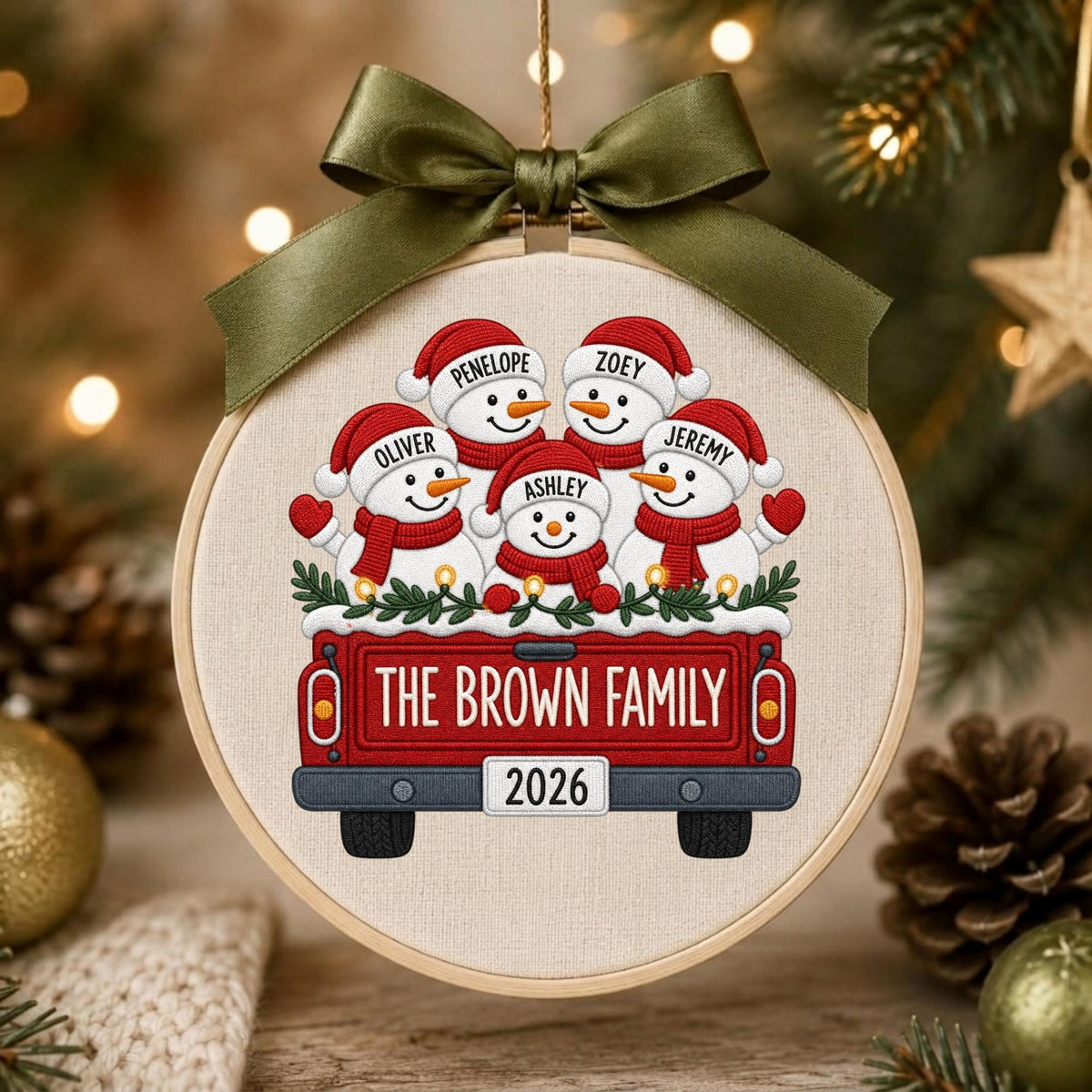 May include: A round embroidery hoop ornament featuring a red truck illustration carrying five snowmen with red hats and scarves. The truck tailgate displays the text THE BROWN FAMILY and the year 2026. An olive green satin ribbon bow is tied at the top.