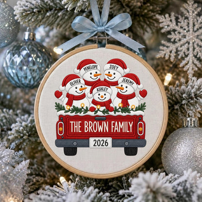 May include: A round wooden embroidery hoop ornament featuring a red truck illustration carrying five snowmen with the names Penelope, Zoey, Oliver, Ashley, and Jeremy. The truck displays the text THE BROWN FAMILY and the year 2026, topped with a light blue ribbon.