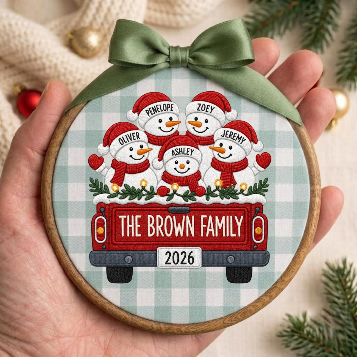 May include: A round wooden hoop ornament featuring a fabric insert with a light green and white buffalo check pattern. The design shows five snowmen in red hats and scarves riding in a red pickup truck with the text THE BROWN FAMILY and 2026.