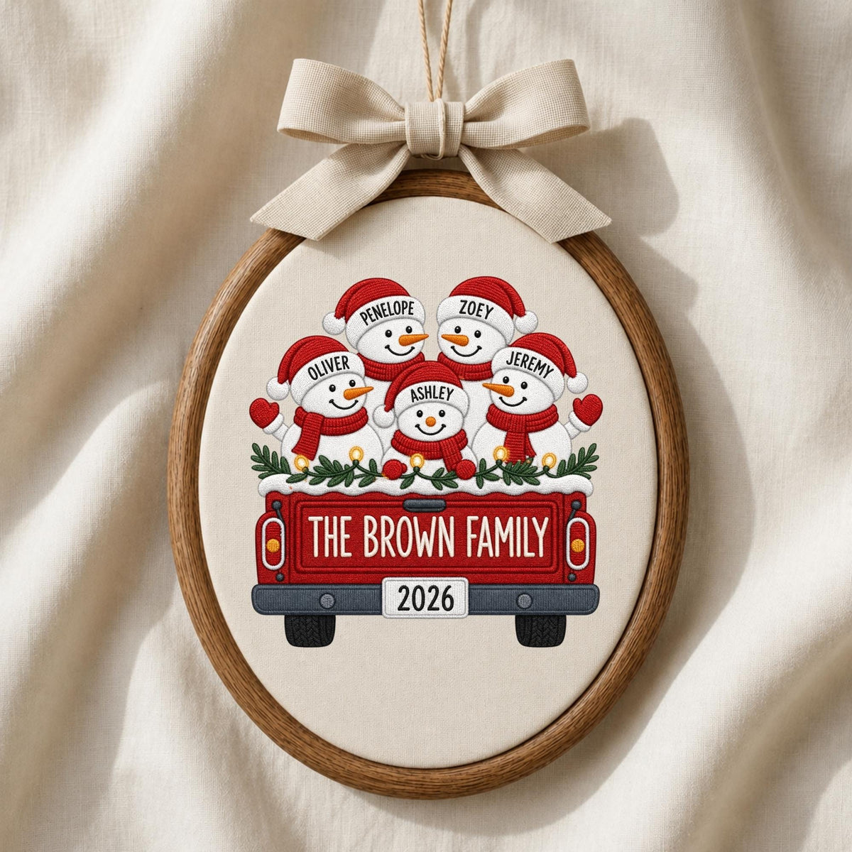 May include: An oval embroidery hoop ornament featuring a red truck carrying five snowmen wearing red hats and scarves. The snowmen are labeled Penelope, Zoey, Oliver, Ashley, and Jeremy. The truck tailgate reads THE BROWN FAMILY and 2026.