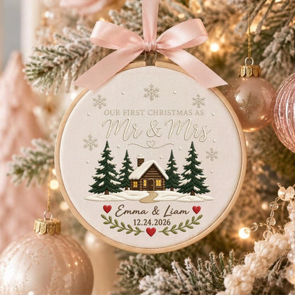 May include: A round wooden embroidery hoop ornament featuring a cream fabric background with a cabin, pine trees, and snowflakes. Text reads: Our First Christmas as Mr &amp; Mrs, Emma &amp; Liam, 12.24.2026. A light pink satin ribbon bow sits on top.