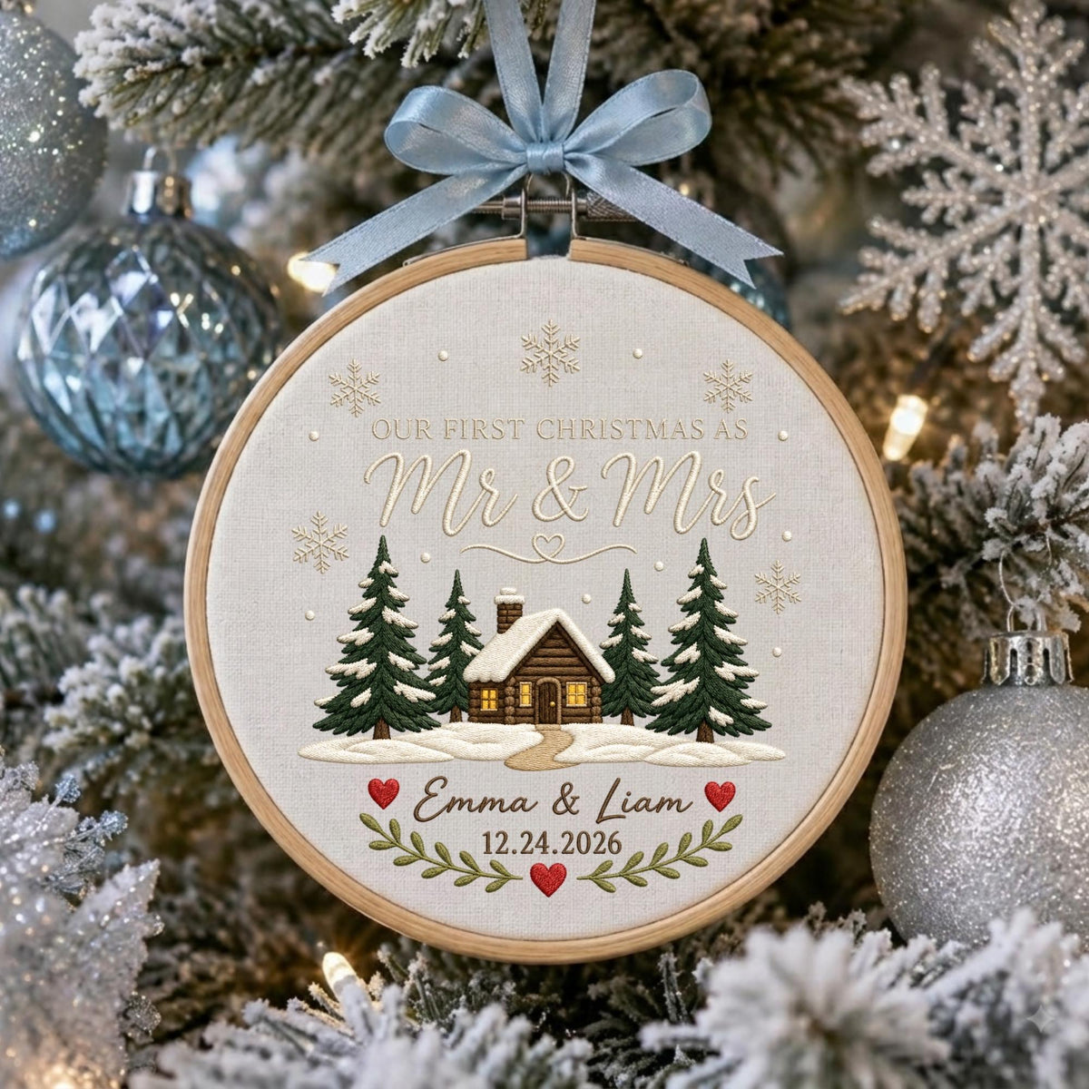 May include: A round wooden embroidery hoop ornament featuring a light fabric background with a stitched cabin, pine trees, and snowflakes. Text reads: Our First Christmas as Mr &amp; Mrs, Emma &amp; Liam, 12.24.2026. A light blue ribbon bow is attached to the top.