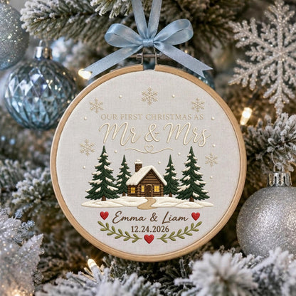 May include: A round wooden embroidery hoop ornament featuring a light fabric background with a stitched cabin, pine trees, and snowflakes. Text reads: Our First Christmas as Mr &amp; Mrs, Emma &amp; Liam, 12.24.2026. A light blue ribbon bow is attached to the top.