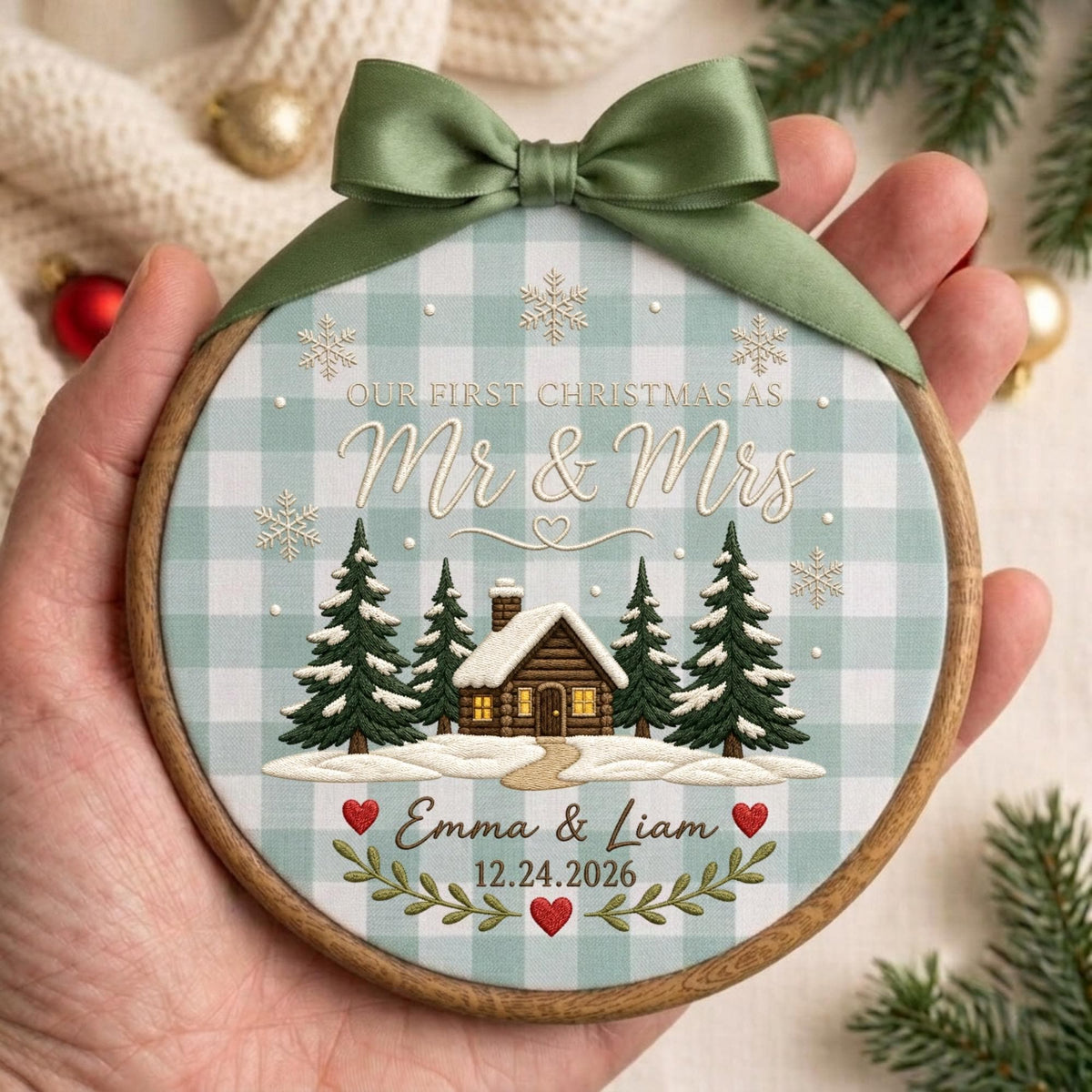 May include: A round wooden embroidery hoop ornament featuring a light blue and white buffalo check fabric background. The design includes a cabin, pine trees, snowflakes, and the text "OUR FIRST CHRISTMAS AS Mr &amp; Mrs Emma &amp; Liam 12.24.2026" with a green satin bow at the top.