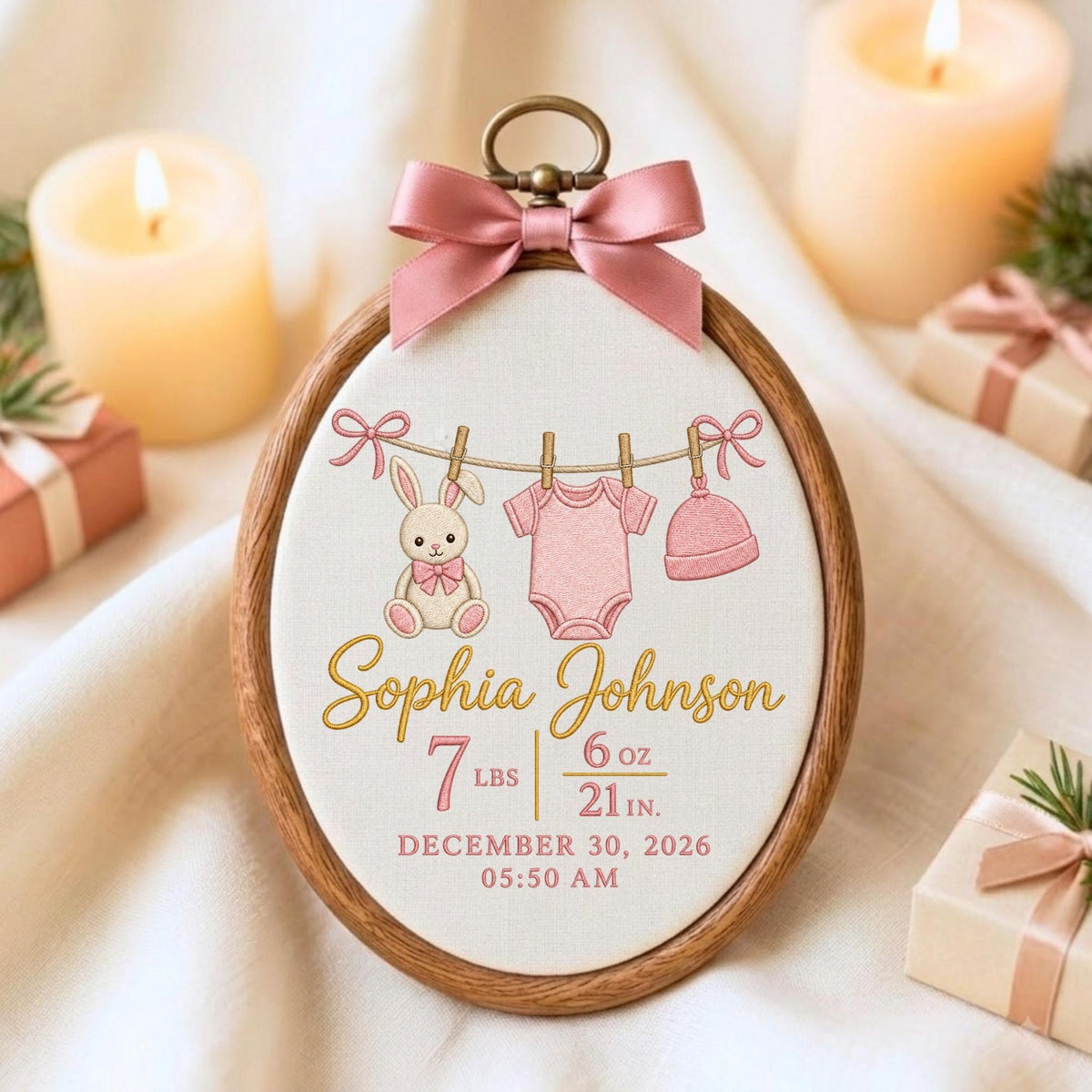 May include: An oval wooden embroidery hoop frame containing a white fabric panel with a pink bow at the top. The design features a rabbit, a baby bodysuit, and a hat hanging on a line, with the text Sophia Johnson, 7 lbs 6 oz, 21 in, December 30, 2026, 05:50 AM.
