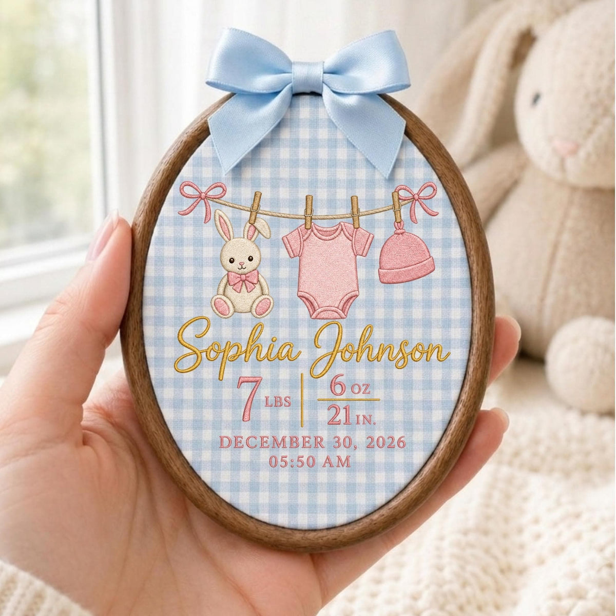 May include: An oval wooden embroidery hoop frame containing fabric with a light blue gingham pattern. It features an embroidered clothesline with a bunny, a pink onesie, and a pink hat. Text reads: Sophia Johnson, 7 lbs 6 oz, 21 in, December 30, 2026, 05:50 AM. A light blue satin bow is attached to the top.