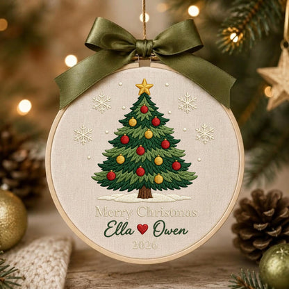 May include: A round embroidery hoop ornament featuring a stitched green Christmas tree with red and yellow ornaments, a gold star topper, and white snowflakes on beige fabric. It includes the text Merry Christmas, Ella, a heart, Owen, and 2026, topped with a dark olive green satin ribbon bow.