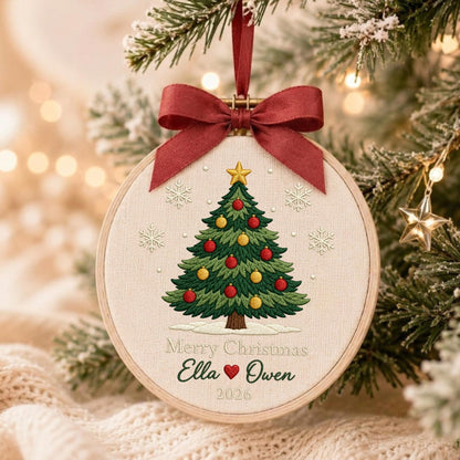 May include: A round wooden embroidery hoop ornament featuring a stitched green Christmas tree with red and yellow ornaments, a gold star topper, and white snowflakes. Below the tree, the text reads Merry Christmas Ella, a red heart, Owen, 2026.