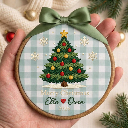 May include: A round wooden embroidery hoop ornament featuring a light blue and white gingham fabric background. The center displays an embroidered green Christmas tree with red and yellow ornaments, a gold star, and snowflakes. Text reads Merry Christmas Ella and Owen 2026, topped with a sage green satin ribbon bow.