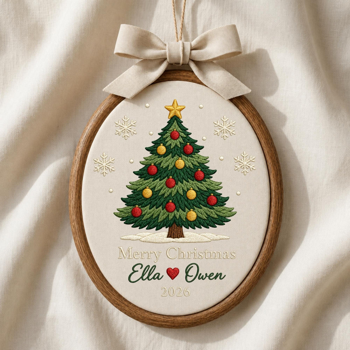 May include: An oval embroidery hoop ornament featuring a green Christmas tree with red and yellow ornaments, a gold star topper, and white snowflakes. The text reads Merry Christmas, Ella, a red heart, Owen, and 2026. A beige ribbon bow is at the top.