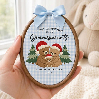 May include: An oval wooden embroidery hoop ornament featuring a light blue and white gingham fabric background. It displays three embroidered brown bears wearing red Santa hats, two green pine trees, and the text First Christmas as my Grandparents, Love from, William 2026. A light blue satin ribbon bow is attached to the top.