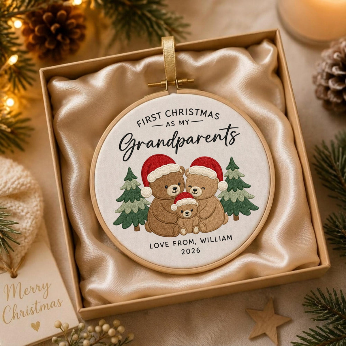 May include: A round embroidery hoop ornament featuring three brown teddy bears wearing red Santa hats, flanked by two green pine trees. The text reads FIRST CHRISTMAS AS MY Grandparents, LOVE FROM, WILLIAM, 2026. The ornament is inside a gift box.