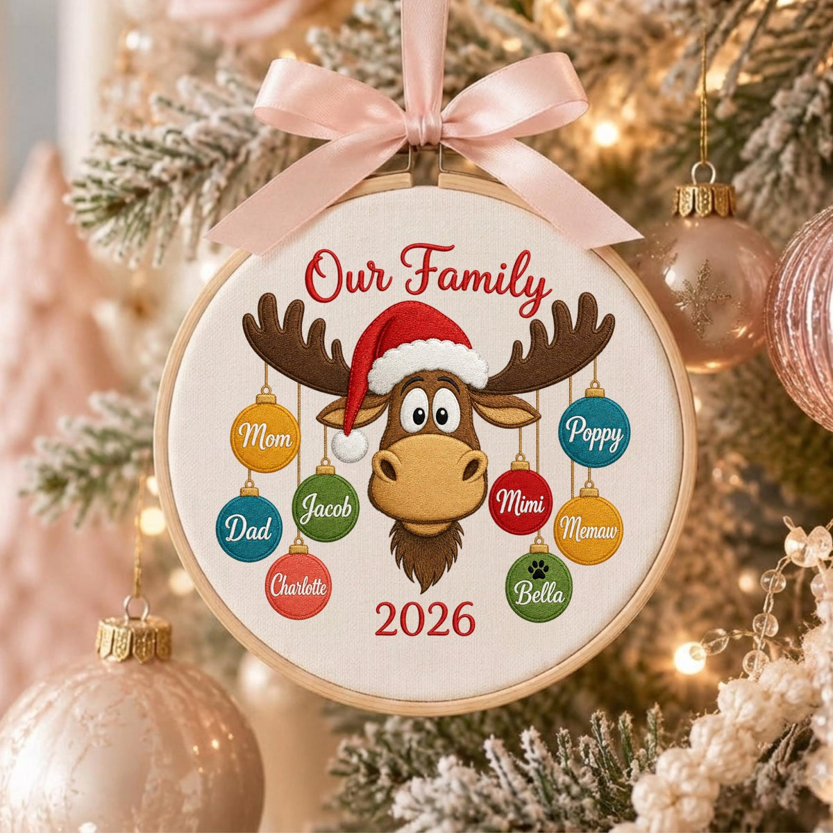 May include: A round wooden embroidery hoop ornament featuring a light-colored fabric center with a brown moose wearing a red Santa hat. The text Our Family and 2026 are embroidered in red. Eight colorful ornaments hang from the moose's antlers.