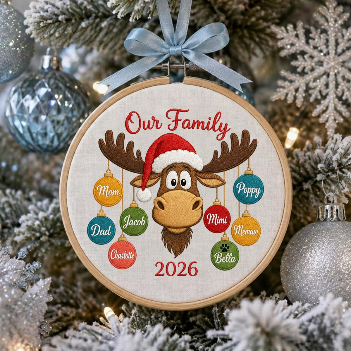 May include: A round wooden embroidery hoop ornament featuring a light-colored fabric center with an embroidered brown moose wearing a red Santa hat. The text Our Family and 2026 are stitched in red. Colorful ornaments hang from the moose antlers.
