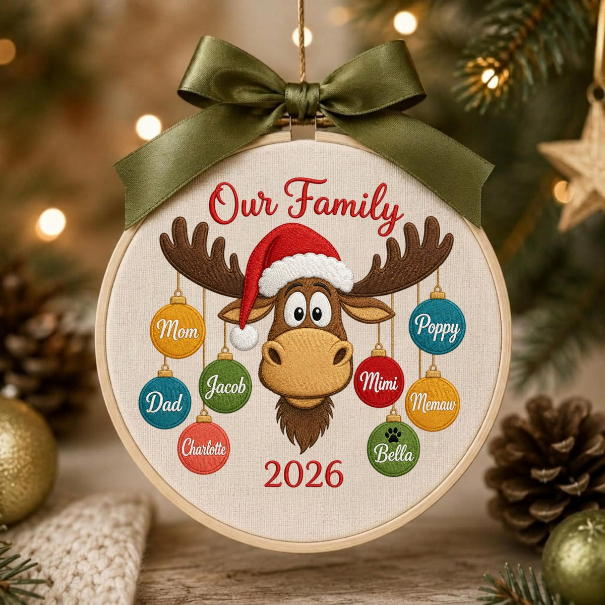 May include: A round embroidery hoop ornament featuring a brown moose wearing a red Santa hat. The text Our Family and 2026 are embroidered in red. Eight colorful ornaments hang from the moose antlers, each labeled with names: Mom, Dad, Charlotte, Jacob, Mimi, Memaw, Poppy, and Bella. An olive green ribbon bow is tied at the top.