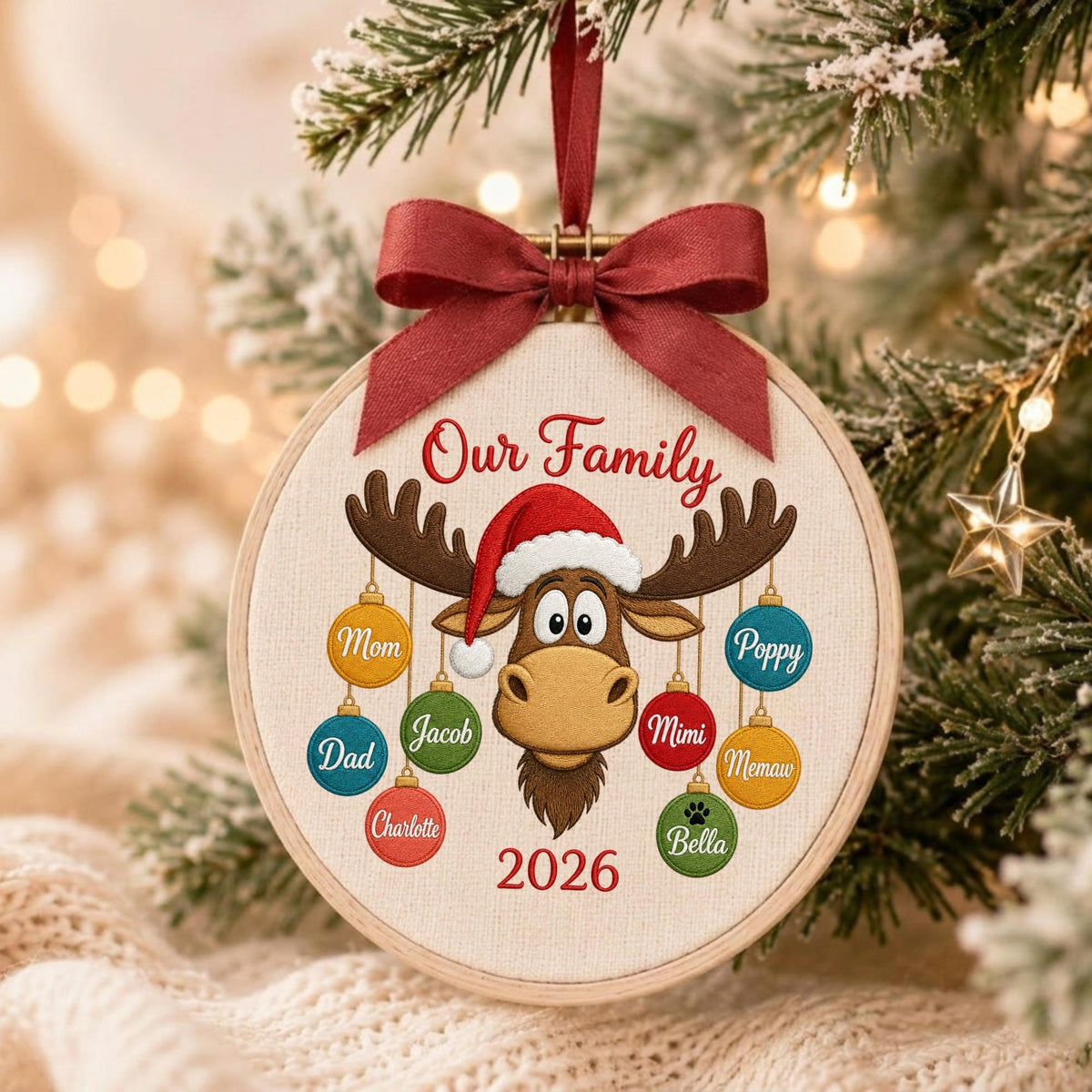 May include: A round wooden embroidery hoop ornament featuring a beige fabric center with the text Our Family and 2026. A cartoon moose wearing a Santa hat is centered, surrounded by colorful hanging ornaments labeled Mom, Poppy, Dad, Jacob, Mimi, Charlotte, Memaw, and Bella.