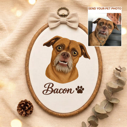May include: An oval wooden embroidery hoop containing a fabric portrait of a brown dog with a gray muzzle and visible teeth. The name Bacon is embroidered in brown script below the dog, followed by a paw print. A beige bow sits at the top.