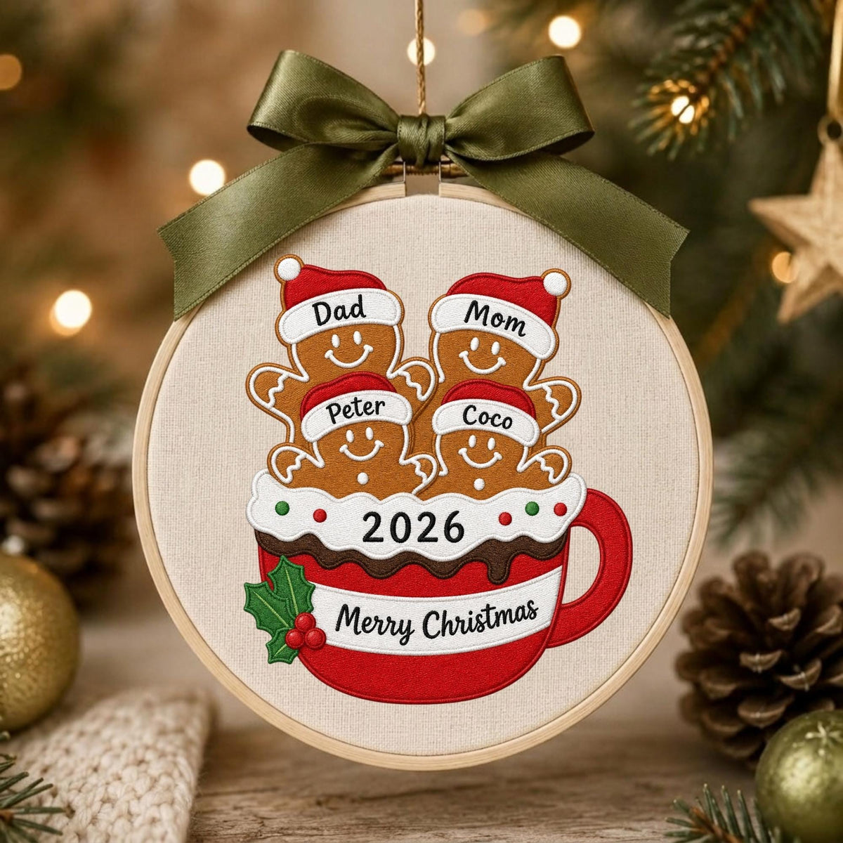 May include: A round embroidery hoop ornament featuring a red mug filled with four gingerbread people wearing Santa hats labeled Dad, Mom, Peter, and Coco. The mug displays the text 2026 and Merry Christmas, accented by a green ribbon bow on top.