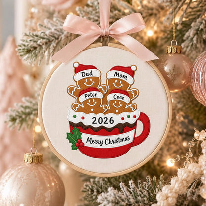 May include: A round wooden embroidery hoop ornament featuring a stitched design of four gingerbread figures in a red mug. The figures wear Santa hats labeled Dad, Mom, Peter, and Coco. The mug displays 2026 and Merry Christmas with a pink ribbon.