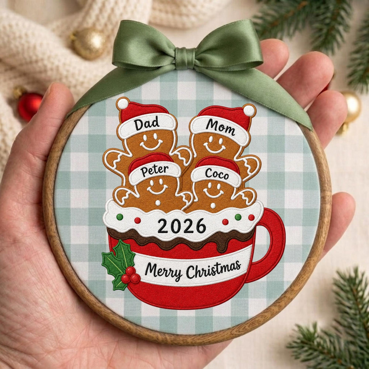 May include: A round wooden embroidery hoop ornament featuring a light blue and white gingham fabric background. It displays four gingerbread figures in a red mug with the text 2026 and Merry Christmas, topped with a green satin ribbon bow.