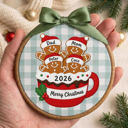 May include: A round wooden embroidery hoop ornament featuring a light blue and white gingham fabric background. It displays four gingerbread figures in a red mug with the text 2026 and Merry Christmas, topped with a green satin ribbon bow.
