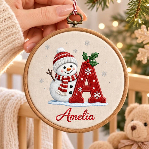 May include: A circular wooden embroidery hoop containing a fabric panel with a stitched design of a snowman wearing a red and white striped hat and scarf, a large red letter A decorated with snowflakes and holly, and the name Amelia in red script.