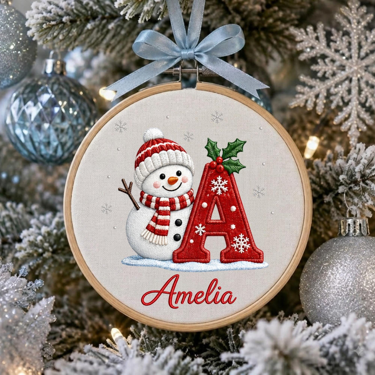 May include: A circular wooden embroidery hoop ornament featuring a white fabric background. The design includes an embroidered snowman wearing a red and white striped hat and scarf, a large red letter A with holly, and the name Amelia in red script.
