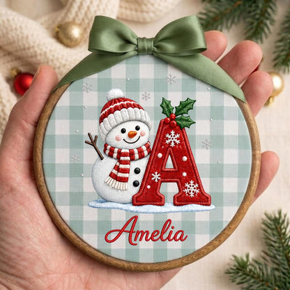 May include: A round wooden embroidery hoop ornament featuring a light green and white gingham fabric background. The design includes a snowman wearing a red and white striped hat and scarf, a large red letter A with holly, and the name Amelia in red.