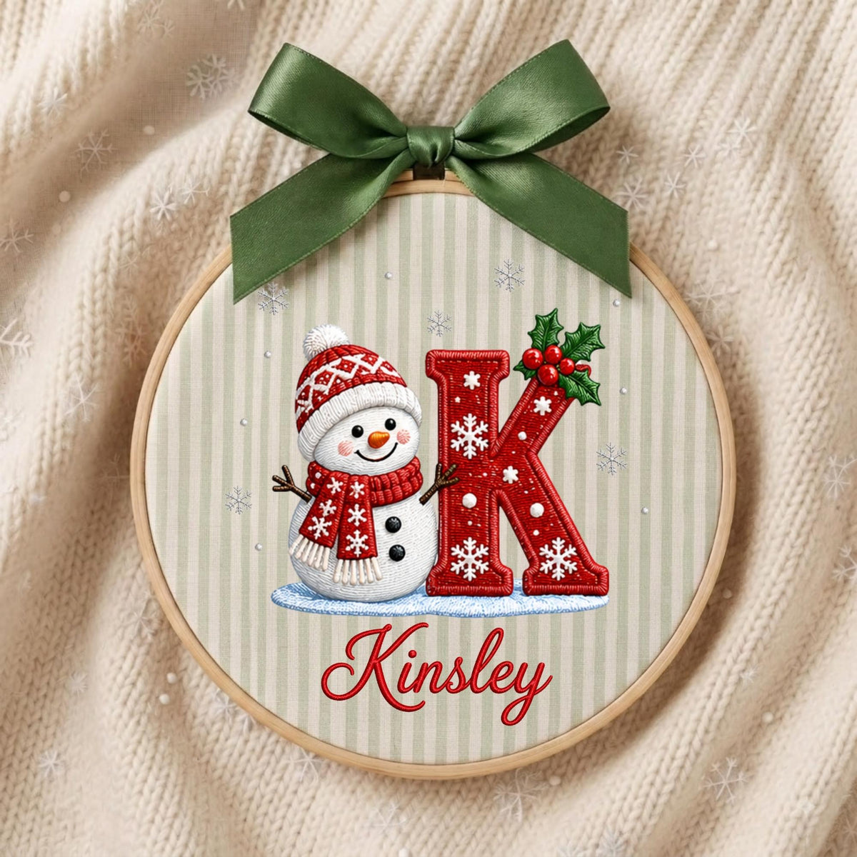May include: A circular embroidery hoop featuring a light beige striped fabric with a snowman wearing a red patterned hat and scarf, a large red letter K decorated with white snowflakes and holly, and the name Kinsley in red script, topped with a green bow.
