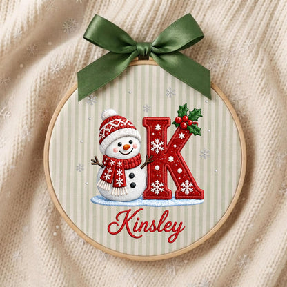 May include: A circular embroidery hoop featuring a light beige striped fabric with a snowman wearing a red patterned hat and scarf, a large red letter K decorated with white snowflakes and holly, and the name Kinsley in red script, topped with a green bow.