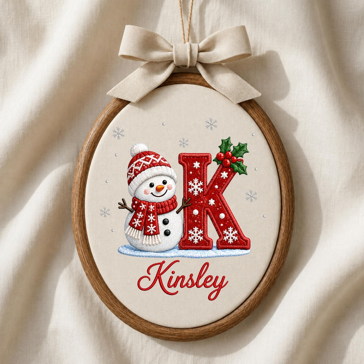 May include: An oval wooden embroidery hoop containing fabric with a stitched snowman wearing a red and white patterned hat and scarf, a large red letter K decorated with white snowflakes and holly, and the name Kinsley in red script text.