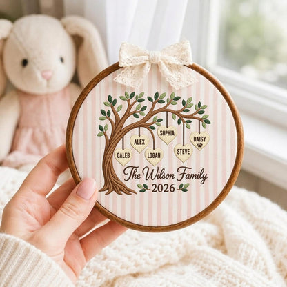 May include: A circular wooden embroidery hoop frame containing fabric with a light pink and white vertical striped pattern. An embroidered brown tree features hanging heart-shaped ornaments labeled with the names Caleb, Alex, Logan, Sophia, Steve, and Daisy, with the text The Wilson Family 2026 below. A cream-colored lace bow is attached to the top.
