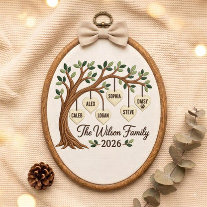 May include: An oval embroidery hoop containing a fabric panel with a stitched tree design. Hanging from the branches are six heart-shaped tags with the names Caleb, Alex, Logan, Sophia, Steve, and Daisy. Text reads The Wilson Family 2026.