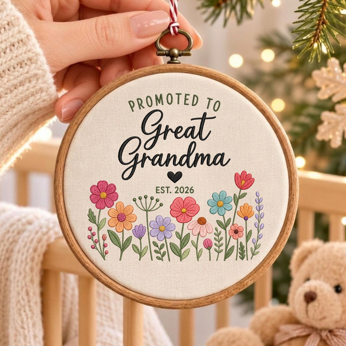 May include: A round wooden embroidery hoop containing off-white fabric with the embroidered text "PROMOTED TO Great Grandma" and "EST. 2026" above a colorful floral arrangement featuring pink, orange, blue, and purple flowers with green stems.