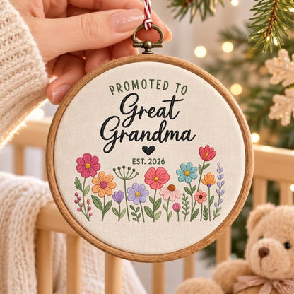 May include: A round wooden embroidery hoop containing off-white fabric with the embroidered text "PROMOTED TO Great Grandma" and "EST. 2026" above a colorful floral arrangement featuring pink, orange, blue, and purple flowers with green stems.