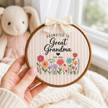 May include: A circular wooden embroidery hoop frame containing fabric with vertical light pink stripes. The text reads PROMOTED TO Great Grandma EST. 2026 with a black heart. Below is a colorful floral illustration. A cream lace bow is attached.