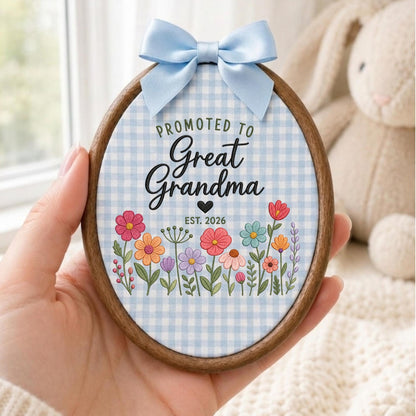May include: An oval wooden embroidery hoop frame containing fabric with a light blue gingham pattern. The text reads PROMOTED TO Great Grandma EST. 2026 above a colorful illustration of various wildflowers. A light blue satin bow is attached at the top.