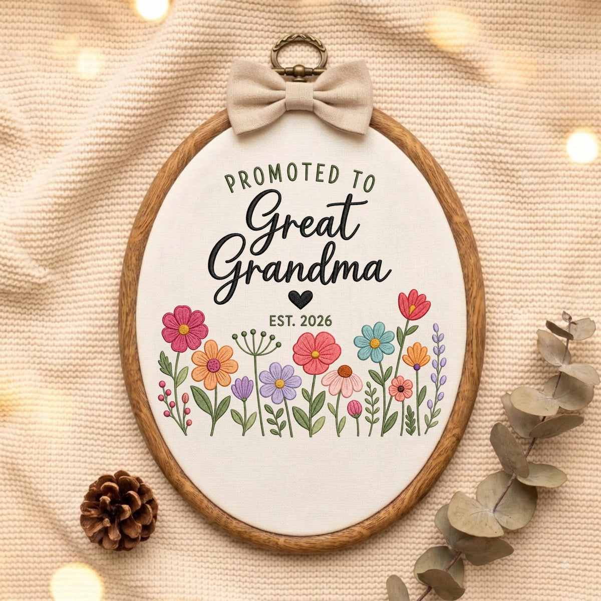 May include: An oval wooden embroidery hoop containing white fabric with the embroidered text Promoted to Great Grandma Est. 2026, a small black heart, and a colorful floral arrangement. A beige fabric bow is attached to the top metal hanger.