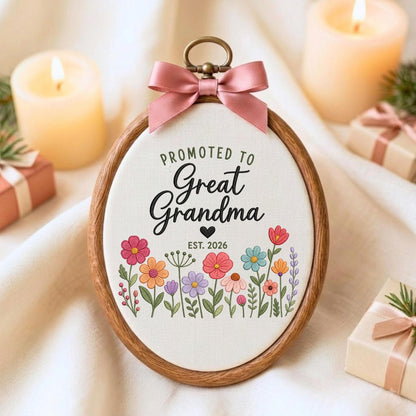 May include: An oval wooden embroidery hoop containing white fabric with the text "PROMOTED TO Great Grandma" and "EST. 2026" in black lettering, accented by a small black heart and a colorful floral illustration. A dusty pink satin bow is attached.