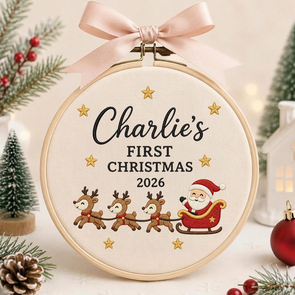 May include: A round wooden embroidery hoop containing cream-colored fabric with the embroidered text Charlie's FIRST CHRISTMAS 2026, surrounded by gold stars, three reindeer, and Santa in a red sleigh, topped with a light pink satin ribbon bow.