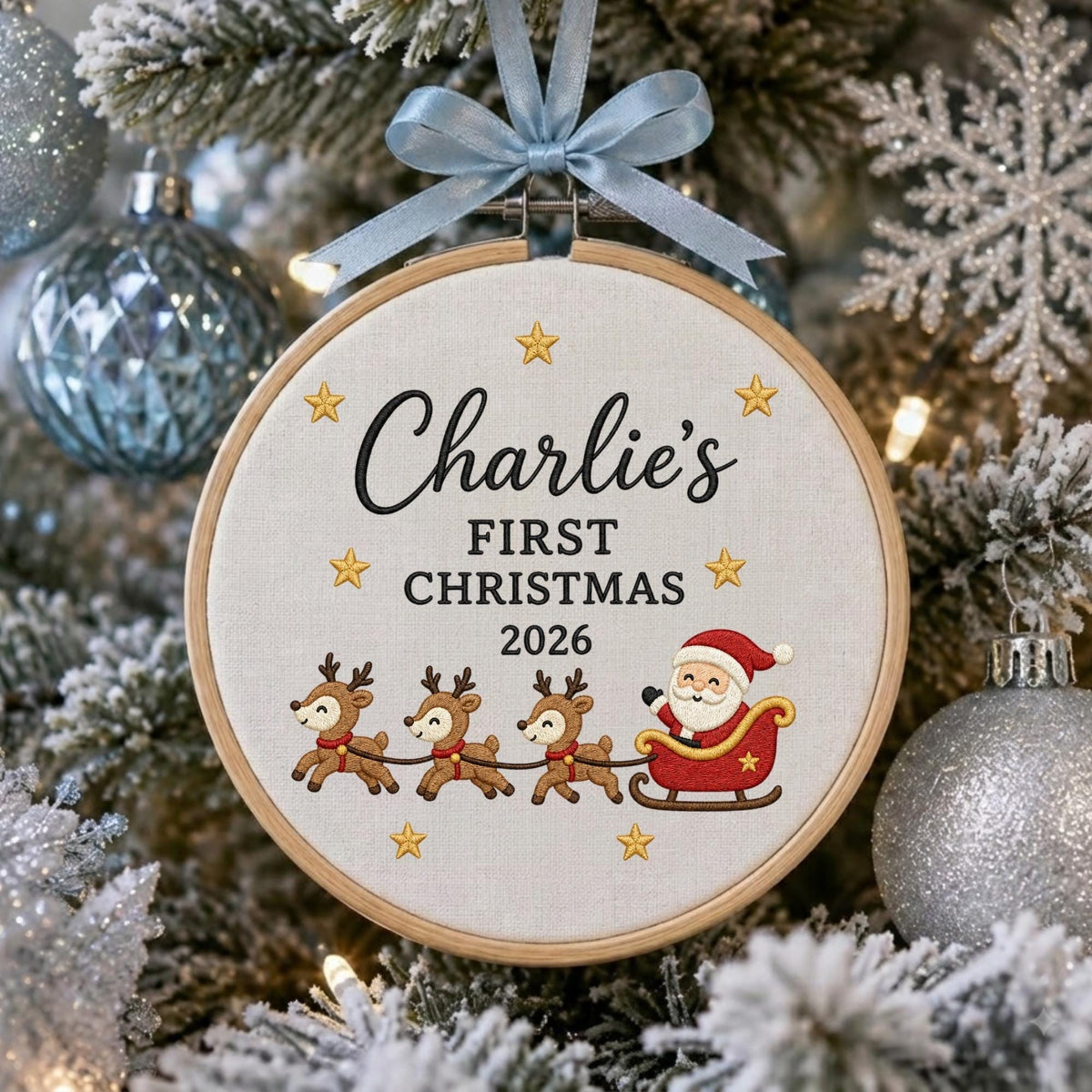 May include: A round wooden embroidery hoop ornament featuring white fabric with embroidered text Charlie's FIRST CHRISTMAS 2026, yellow stars, three reindeer, and Santa Claus in a red sleigh, topped with a light blue satin ribbon bow.