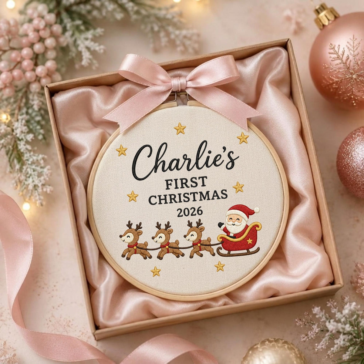 May include: A round wooden embroidery hoop containing cream-colored fabric with the embroidered text Charlie's FIRST CHRISTMAS 2026, surrounded by gold stars and a graphic of Santa Claus in a sleigh pulled by three reindeer, topped with a pink ribbon.