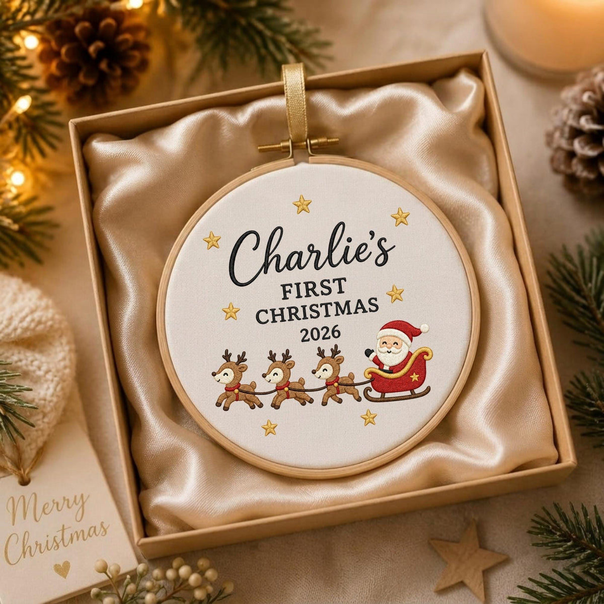 May include: A round wooden embroidery hoop ornament featuring white fabric with the embroidered text Charlie's FIRST CHRISTMAS 2026, surrounded by gold stars and a graphic of Santa Claus in a sleigh pulled by three reindeer, inside a gift box.