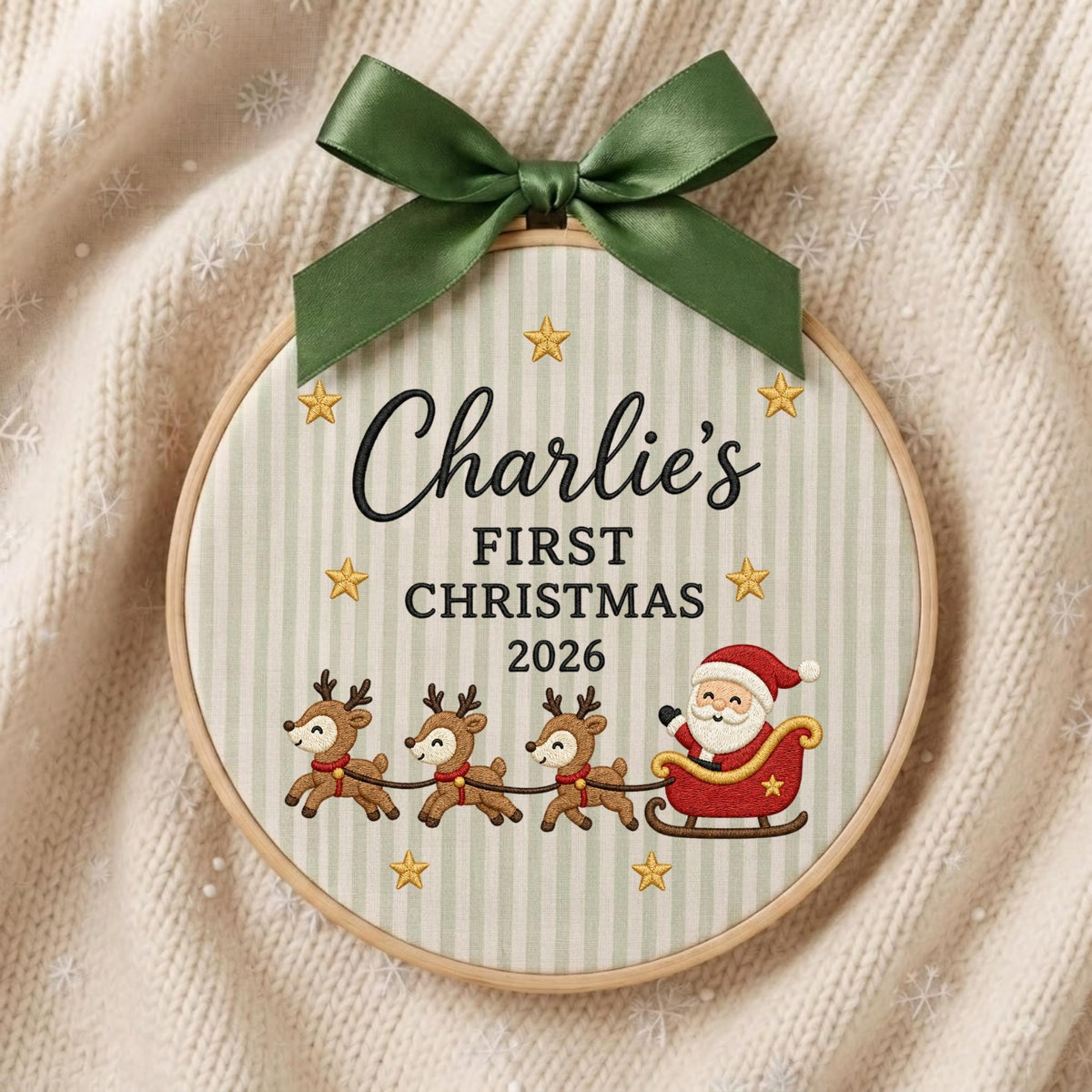 May include: A circular wooden embroidery hoop containing fabric with light green vertical stripes. The text reads Charlie's FIRST CHRISTMAS 2026. Embroidered designs include gold stars, three reindeer, and Santa Claus in a red sleigh, topped with a green ribbon.