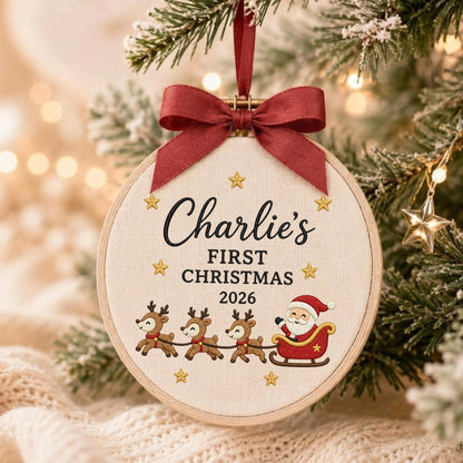 May include: A round wooden embroidery hoop ornament featuring cream fabric with the embroidered text Charlie's FIRST CHRISTMAS 2026, gold stars, three reindeer, and Santa in a red sleigh. A dark red satin ribbon bow is tied at the top for hanging.