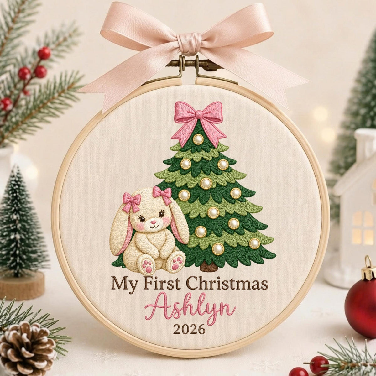 May include: A round wooden embroidery hoop featuring a cream-colored fabric with an embroidered green Christmas tree decorated with white pearls and a pink bow, a cream bunny with pink bows, and the text My First Christmas Ashlyn 2026.