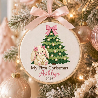 May include: A round wooden embroidery hoop ornament featuring a cream fabric background with an embroidered green Christmas tree decorated with white pearls and a pink bow. A cream bunny with pink bows sits beside the tree. Text reads My First Christmas Ashlyn 2026.