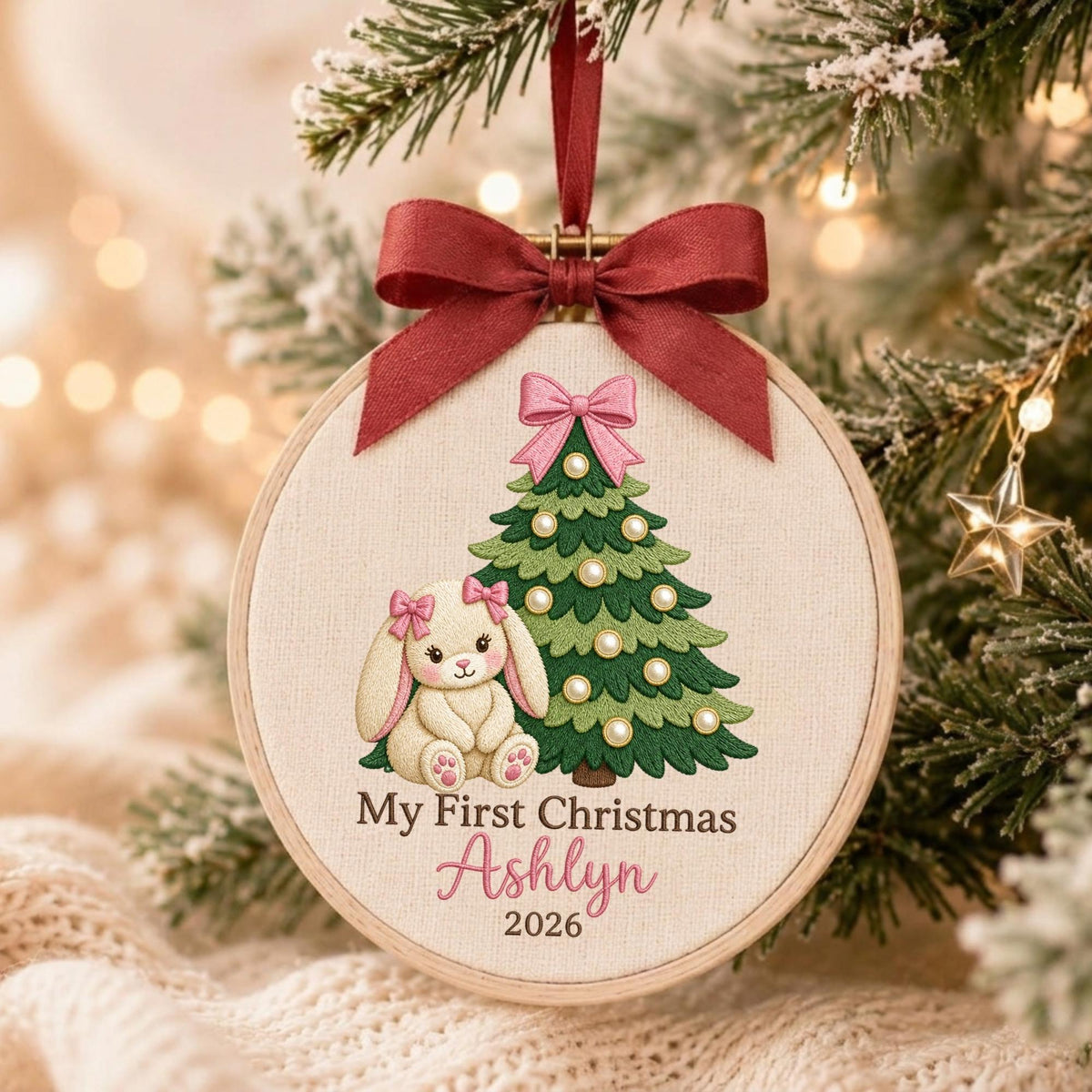 May include: A round wooden embroidery hoop ornament featuring a cream-colored fabric background. The design includes a green Christmas tree with pearl-like ornaments, a pink bow, and a cream-colored bunny with pink bows. Text reads My First Christmas Ashlyn 2026.